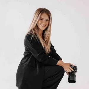 Porträt einer lächelnden Fotografin mit langen blonden Haaren, die in schwarzer Kleidung posiert und eine Kamera in der Hand hält, vor hellem, neutralem Hintergrund.