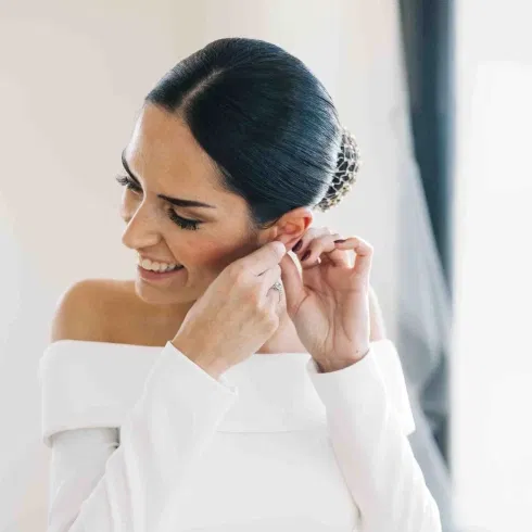 Braut steckt sich einen Ohrring an, während sie in ihrem eleganten, schulterfreien Brautkleid lächelt.