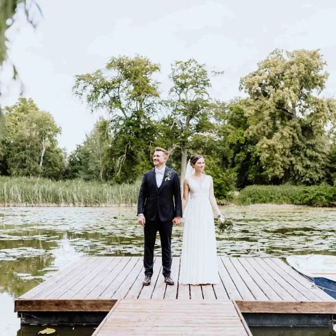 Braut und Bräutigam stehen Hand in Hand auf einem Holzsteg am See, umgeben von Wasserlilien und grüner Natur.