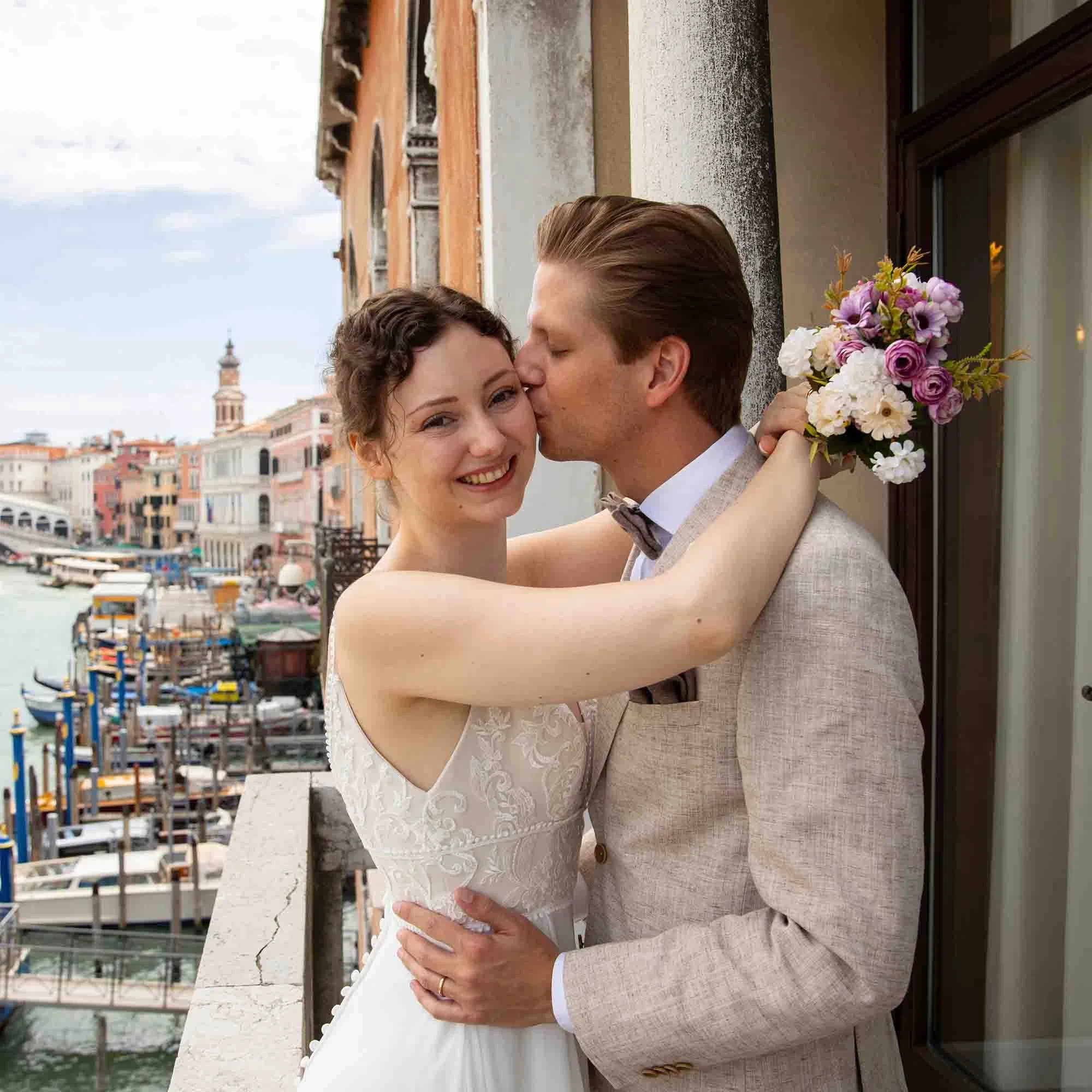 Braut und Bräutigam umarmen sich auf einem Balkon in Venedig, während der Bräutigam die Braut liebevoll auf die Wange küsst.