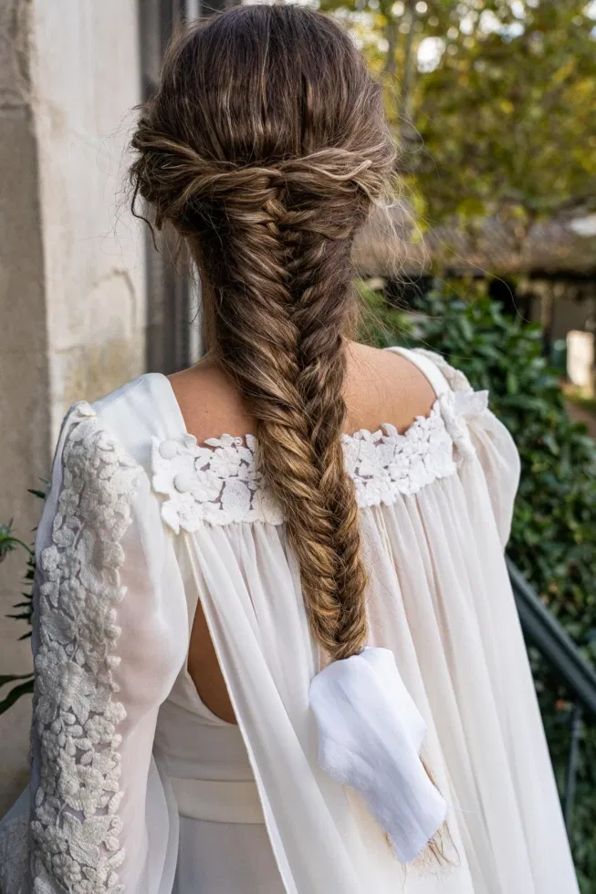 Rückansicht einer Braut mit langem, locker geflochtenem Fischgrätenzopf und einem cremefarbenen Brautkleid mit floraler Spitzenverzierung an den Schultern.