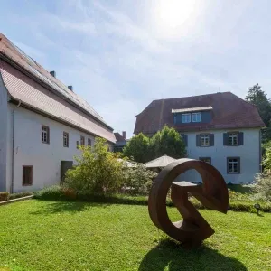 Außenansicht eines historischen Gebäudekomplexes mit zwei Häusern, gepflegtem Garten und moderner Metallskulptur auf der Rasenfläche bei strahlendem Sonnenschein.