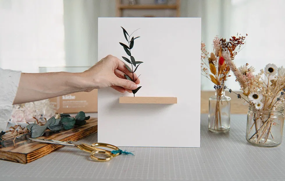 Hand arrangiert einen Eukalyptuszweig auf einem weißen DIY-Bilderrahmen mit kleiner Holzleiste, umgeben von getrockneten Blumen, Schere und Bastelmaterialien.