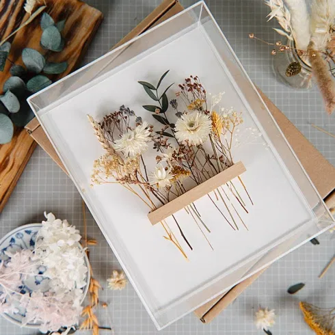 Dekorativer DIY-Bilderrahmen mit zarten getrockneten Blumen in Naturtönen auf weißem Hintergrund, umgeben von Bastelmaterialien und Werkzeugen.