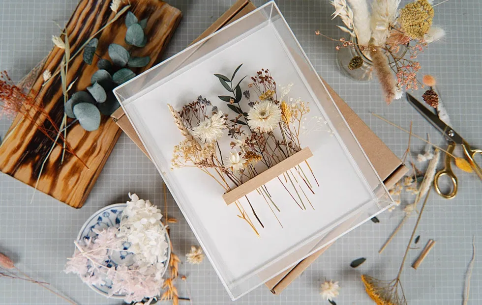 Dekorativer DIY-Bilderrahmen mit zarten getrockneten Blumen in Naturtönen auf weißem Hintergrund, umgeben von Bastelmaterialien und Werkzeugen.