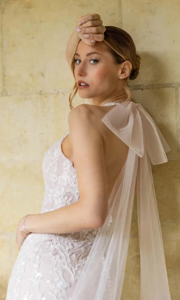 Rückenansicht eines Brautmodells in einem figurbetonten Brautkleid mit zarter Spitze, tiefem Rückenausschnitt und transparenter Tüllschleife am Hals, elegant inszeniert vor einer hellen Steinwand im warmen Licht.