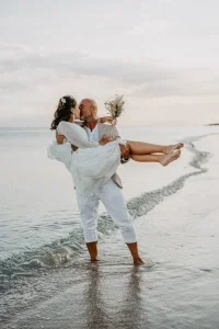 Brautpaar küsst sich am Strand, während der Bräutigam die Braut im weißen Kleid im Arm hält.