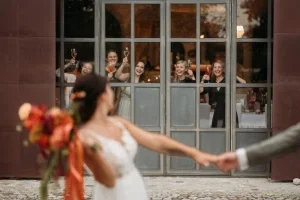 Freunde der Braut stoßen lachend hinter einer großen Fensterscheibe mit Aperitifs auf das Brautpaar an.