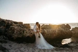 Brautpaar umarmt sich innig zwischen rauen Felsen am Meer und erlebt einen romantischen Sonnenuntergang am Strand.