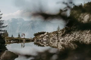 Kleines Brautpaar tanzt inmitten einer beeindruckenden Berglandschaft und spiegelt sich malerisch im stillen See.