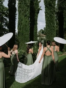 Braut mit langer Schleppe wird von Bridesmaids in grünen Kleidern und weißen Sonnenschirmen durch einen Garten begleitet.