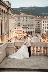 Elegantes Brautpaar umarmt sich auf einer steinernen Terrasse mit Blick über eine historische Stadt bei Abendstimmung.