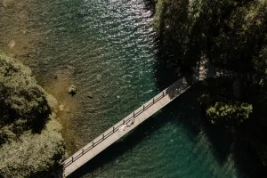 Luftaufnahme eines Brautpaares auf einer Holzbrücke über glasklarem Wasser inmitten dichter Bäume.