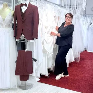 Frau in schwarzer Kleidung steht lächelnd zwischen weißen Brautkleidern in einem Brautmodengeschäft; daneben ein burgunderfarbener Herrenanzug auf einer Schneiderpuppe.