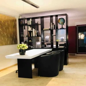 Eleganter Beratungsbereich in einer Goldschmiede mit weißem Tisch, zwei schwarzen Sesseln, dekorativem Bücherregal mit Schmuckvitrinen und einem Blumenarrangement als Tischdeko.