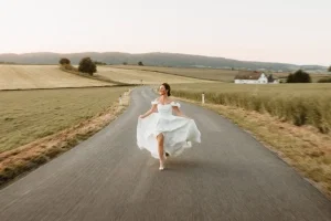 Glückliche Braut läuft in einem schulterfreien Hochzeitskleid über eine kurvige Landstraße durch die Natur.