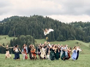 Hochzeitsgesellschaft wirft lachende Braut mit Blumenkranz auf einer grünen Wiese in die Luft.