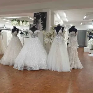 Mehrere opulente Brautkleider mit Spitzendetails und Tüll stehen auf Schneiderpuppen in einem eleganten Brautmodengeschäft, umgeben von Blumenarrangements und heller, moderner Einrichtung.