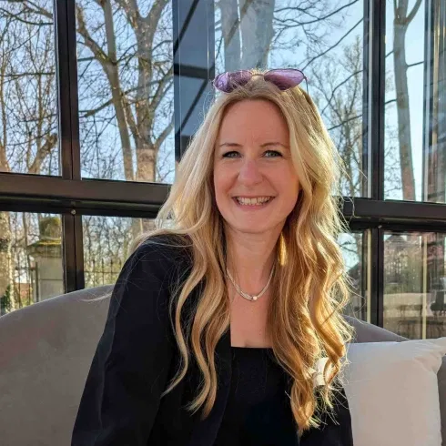 Lächelnde blonde Frau mit langen gewellten Haaren sitzt in einer modernen Glaslounge, Sonnenlicht fällt auf ihr Gesicht.