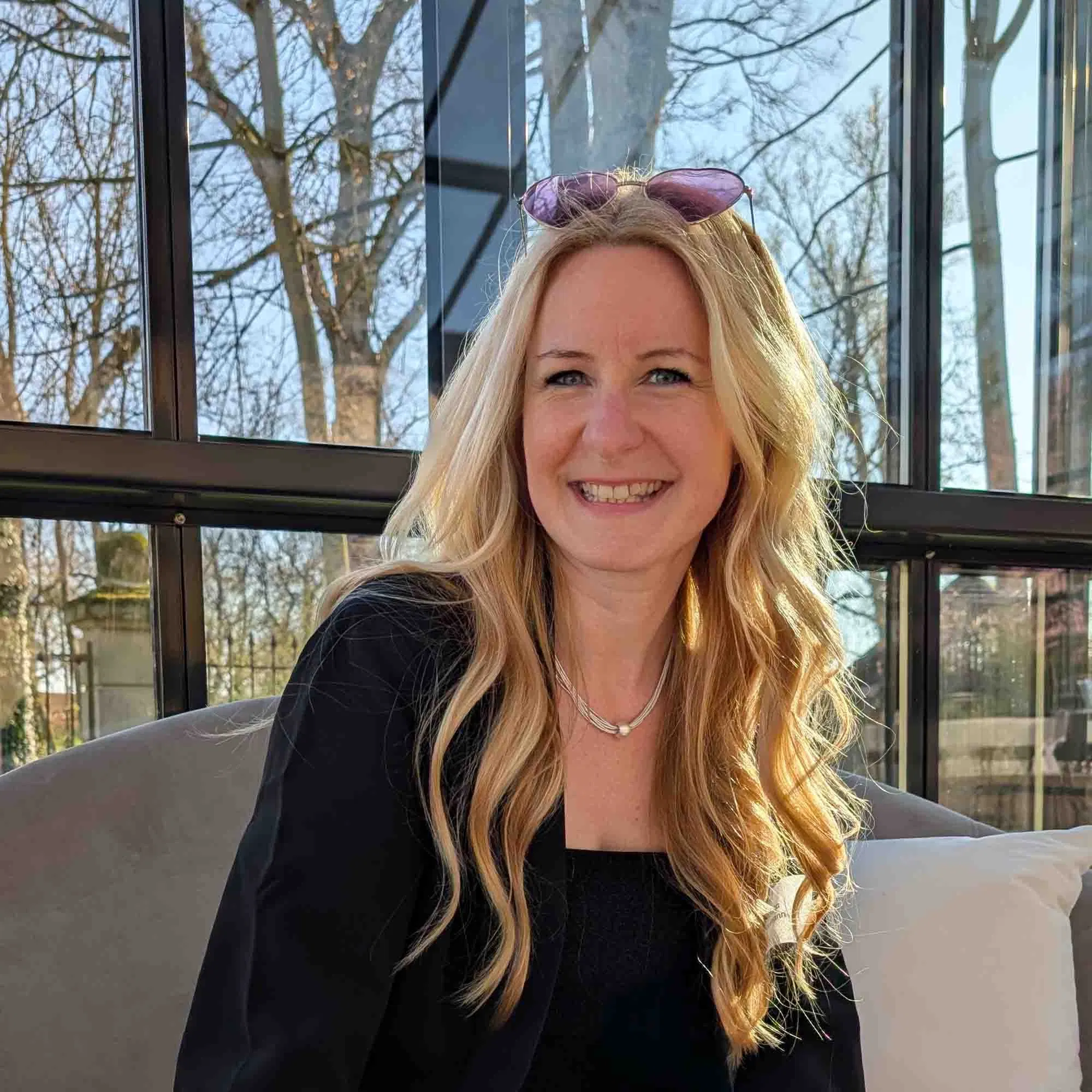 Lächelnde blonde Frau mit langen gewellten Haaren sitzt in einer modernen Glaslounge, Sonnenlicht fällt auf ihr Gesicht.