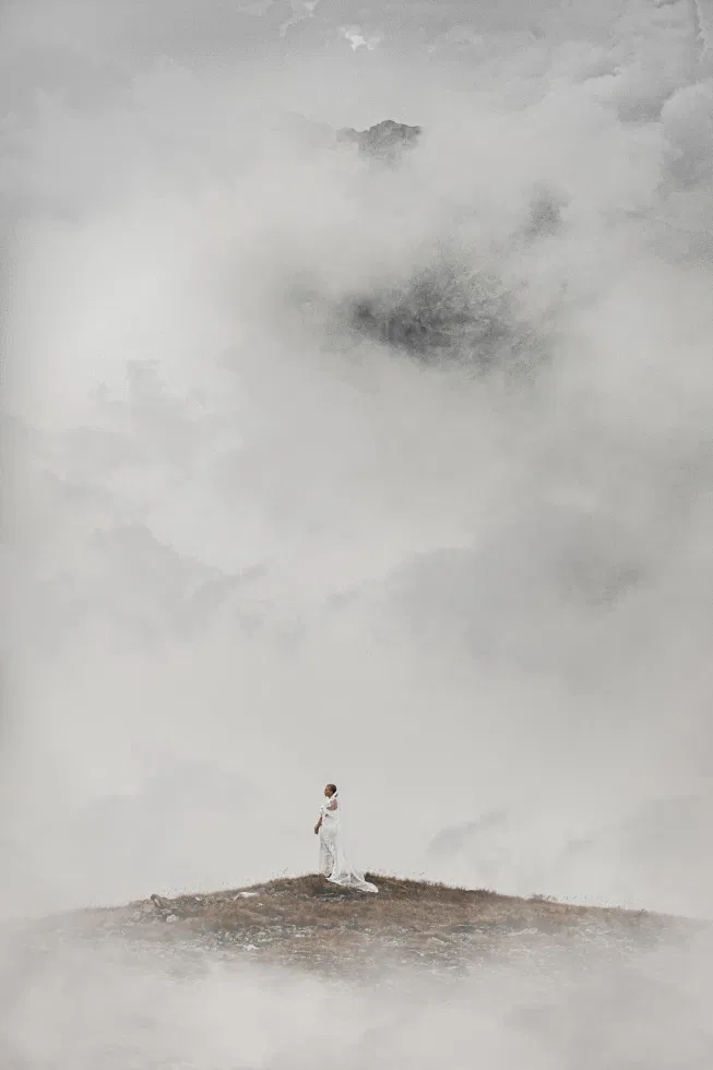 Braut in einem langen weißen Kleid steht allein auf einem nebelverhangenen Berggipfel, umgeben von dichter, mystischer Wolkenlandschaft.