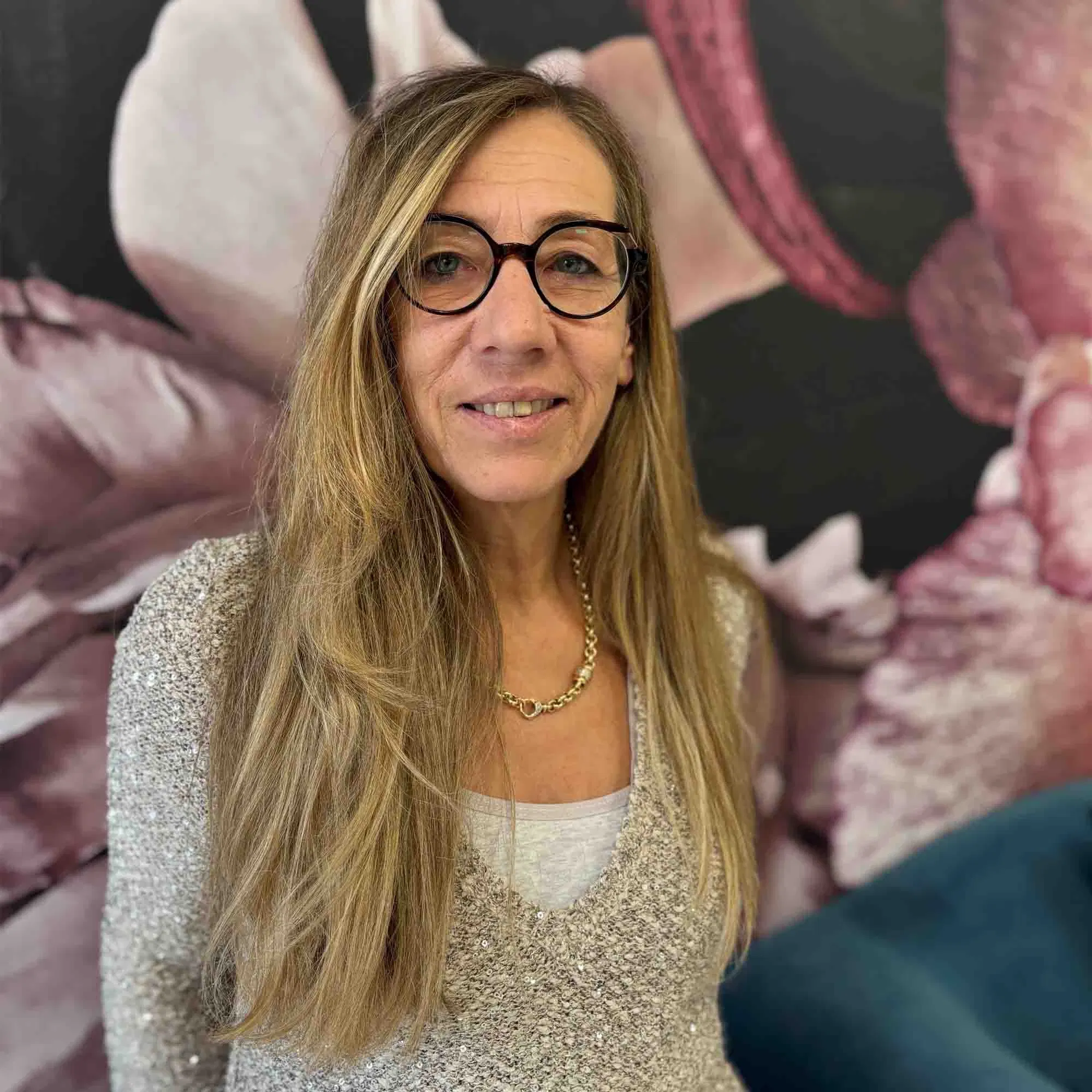 Porträt einer Frau mit langen blonden Haaren und schwarzer Brille, die vor einer floralen, dunkel gehaltenen Wand steht und freundlich in die Kamera lächelt.