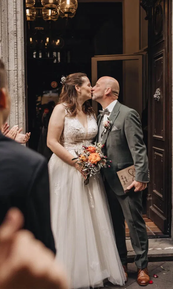 Braut und Bräutigam küssen sich vor der Tür des Standesamts, während Gäste links und rechts applaudieren.