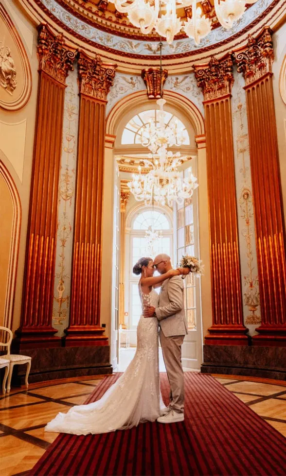 Braut und Bräutigam umarmen sich in einem prunkvollen Saal mit hohen goldenen Säulen und funkelnden Kronleuchtern.