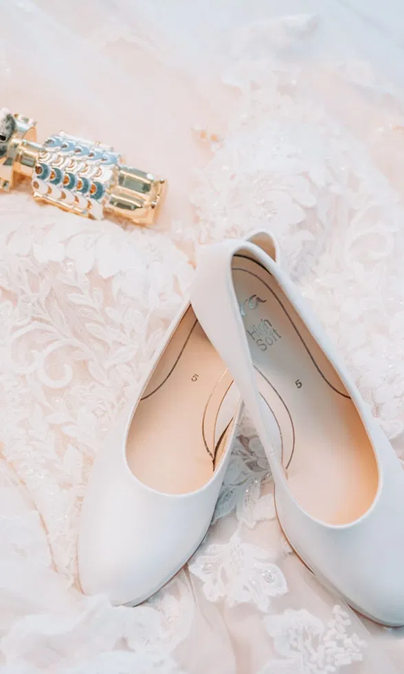 Weiße Brautschuhe liegen auf dem bestickten Spitzenstoff des Brautkleids, daneben ein funkelndes goldenes Accessoire.