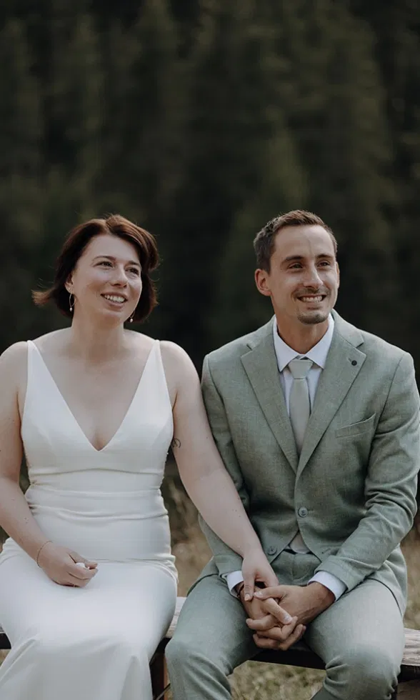 Die Braut und der Bräutigam sitzen Hand in Hand und schauen lächelnd zur Seite, umgeben von Natur.