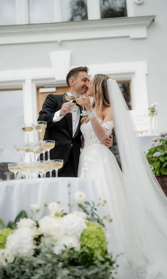 Braut und Bräutigam stoßen mit Champagner an und küssen sich vor dem Hochzeitsturm aus Gläsern.