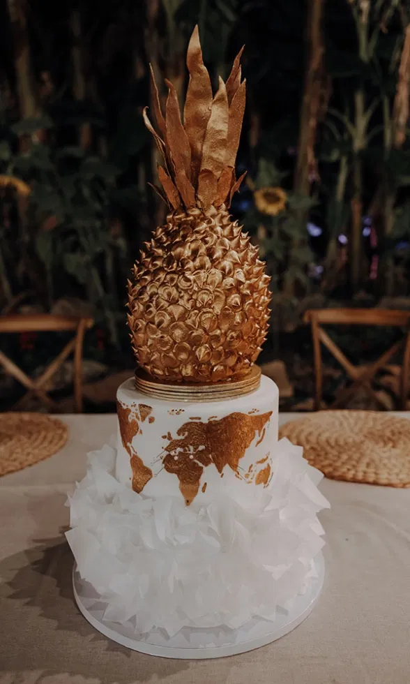 Hochzeitstorte mit goldener Ananas und einer goldenen Weltkarte auf weißem Fondant, verziert mit weißen Blütenrüschen.