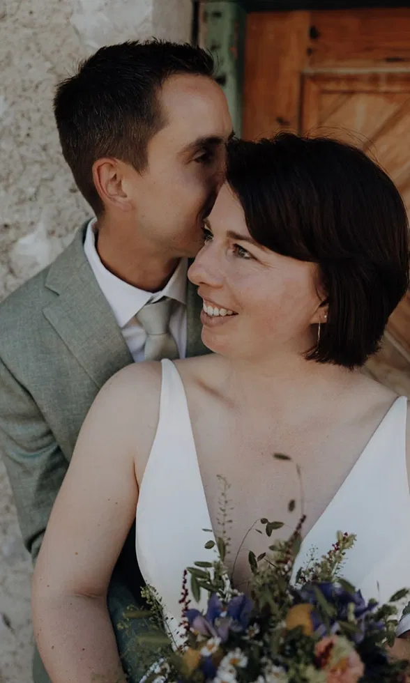 Zärtliches Porträt eines Brautpaares, bei dem der Bräutigam der Braut liebevoll einen Kuss auf die Stirn gibt, während sie lächelnd in die Ferne blickt.