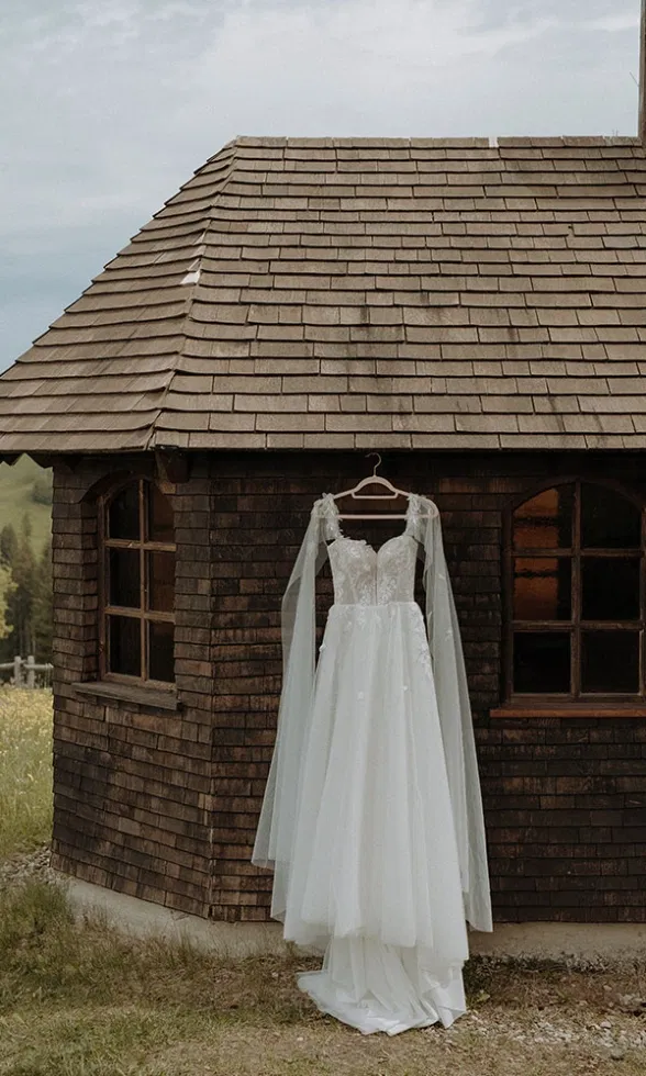 Weißes Brautkleid mit Spitzenoberteil hängt vor einer dunklen Holzhütte und bewegt sich leicht im Wind.