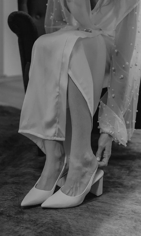 Schwarz-weiß-Aufnahme einer Braut, die sich elegante Pumps anzieht und den Schuhriemen zurechtrückt.