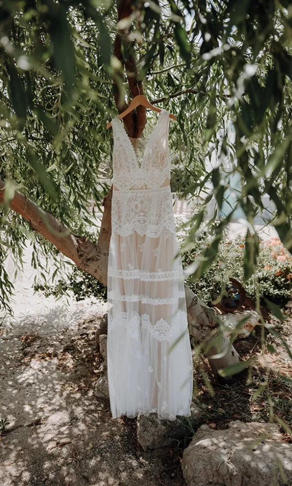 Boho-Brautkleid aus Spitze hängt an einem Ast unter dichtem Grün und wird von natürlichem Licht durch die Blätter umspielt.