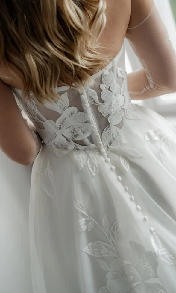 Rückenansicht der Braut mit floraler Spitzenapplikation und Knopfleiste am transparenten Oberteil des Brautkleids.