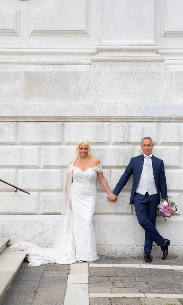 Braut und Bräutigam lehnen entspannt an einer hellen Kirchenwand und halten sich an den Händen, die Braut trägt ein schulterfreies Spitzenbrautkleid mit Schleppe, der Bräutigam einen dunkelblauen Anzug und hält einen Blumenstrauß – ein moderner, ruhiger Hochzeitsmoment.