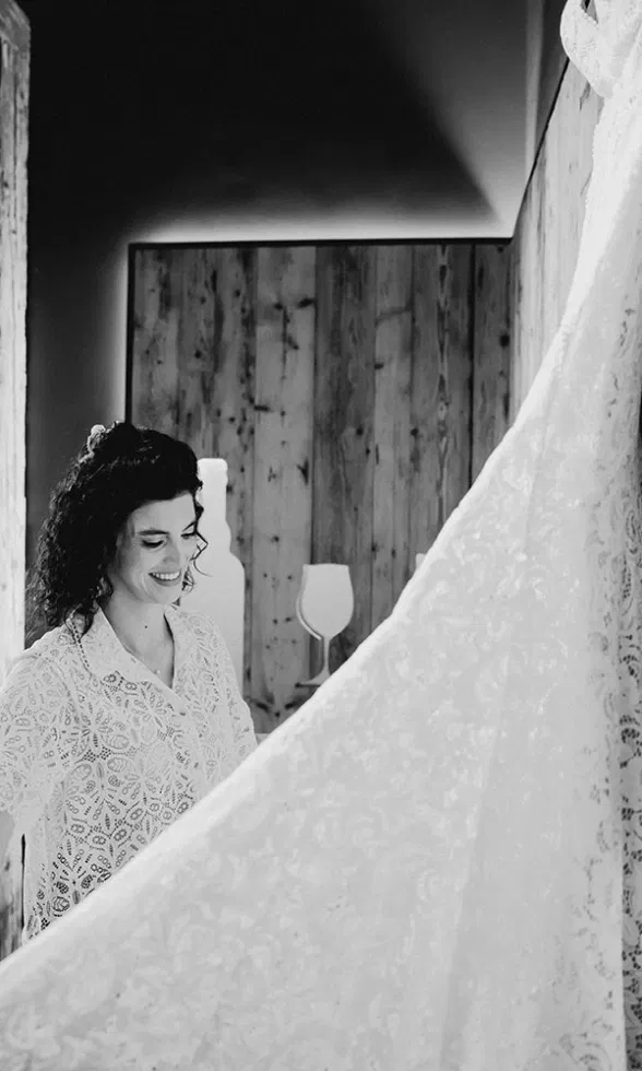 Schwarz-weiß-Aufnahme der Braut beim Getting Ready, sie betrachtet lächelnd ihr Spitzenbrautkleid mit ausladender Schleppe.