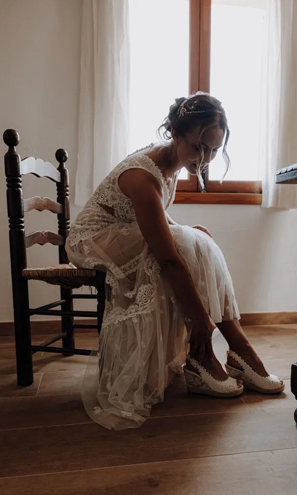 Braut sitzt im Zimmer vor dem Fenster und schlüpft in ihre weißen Wedges, während ihr Spitzenkleid sanft zu Boden fällt.