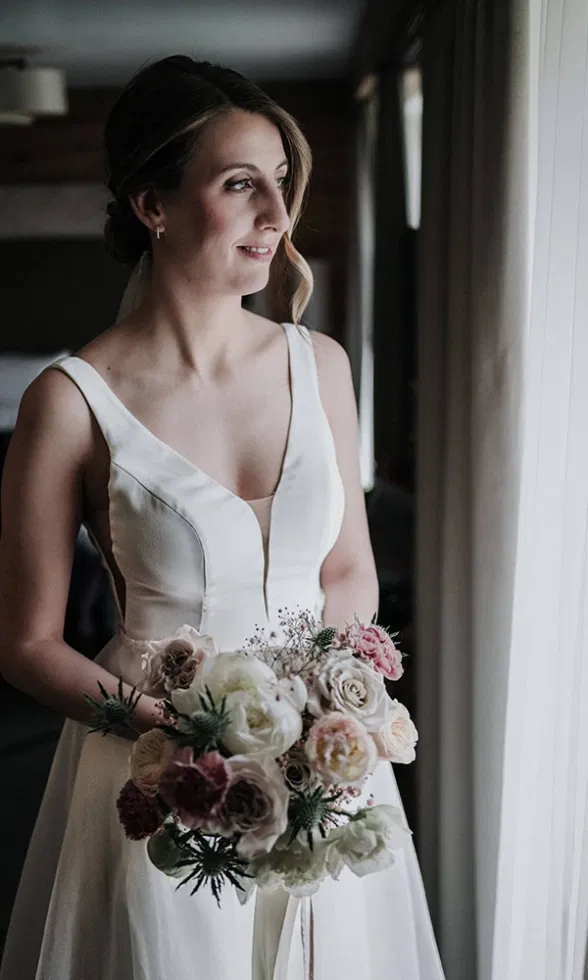 Braut im schlichten Brautkleid steht am Fenster und blickt nach draußen, in den Händen hält sie einen zarten Blumenstrauß in soften Naturtönen.