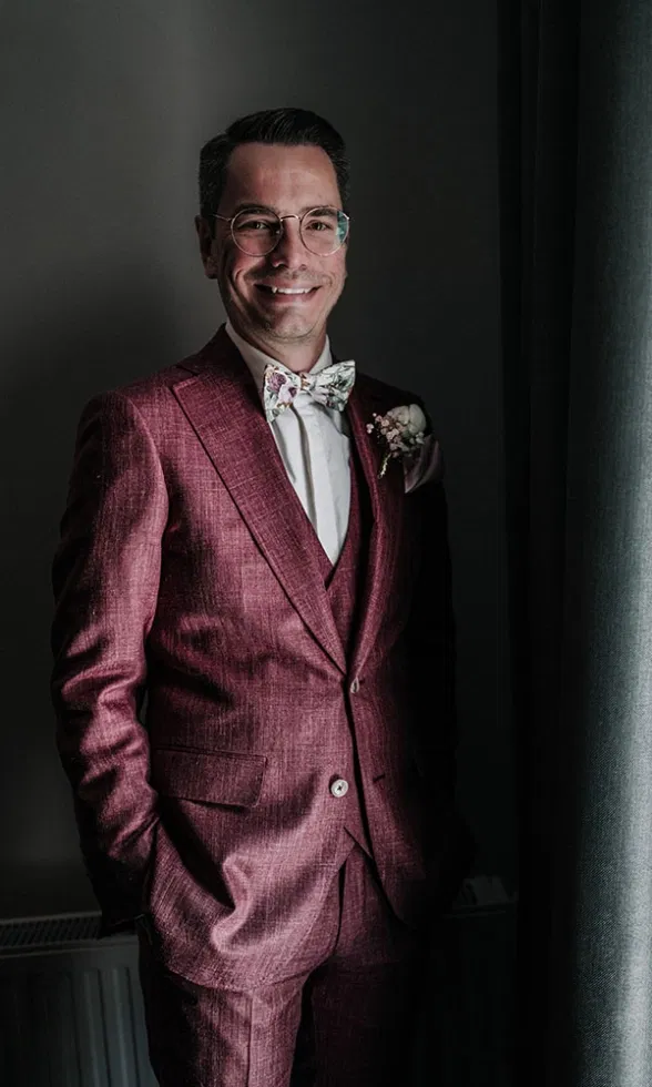 Bräutigam im bordeauxfarbenen Hochzeitsanzug steht am Fenster und lächelt in die Kamera, kombiniert mit floraler Fliege und Ansteckblume.