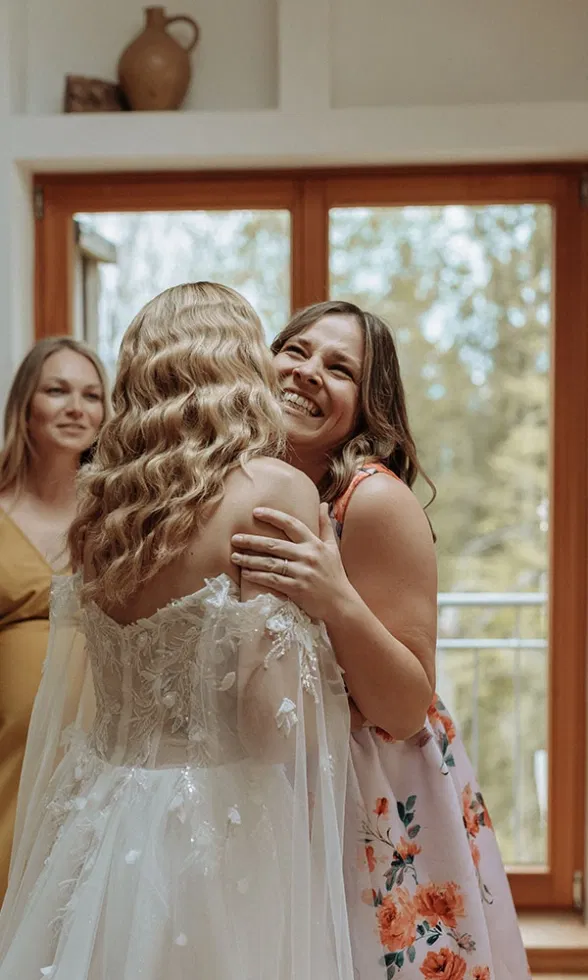 Braut umarmt eine lachende Freundin, während im Hintergrund warmes Licht durch große Fenster fällt.