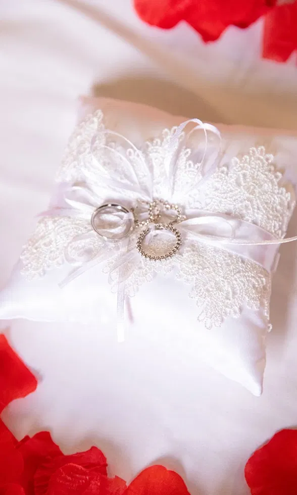 Zwei Trauringe liegen auf einem weißen Ringkissen mit Spitzenapplikation und Schleife, umgeben von roten Rosenblättern – ein detailreicher, romantischer Hochzeitsmoment.