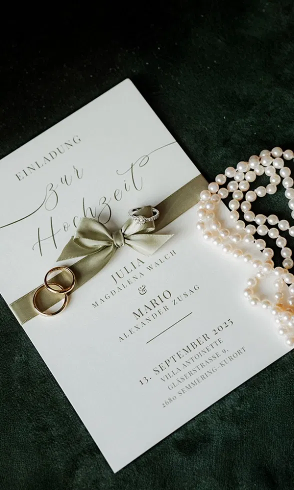 Elegante Hochzeitseinladung mit Schleife, Trauringen und Perlenkette als stilvolles Flatlay auf dunklem Untergrund.