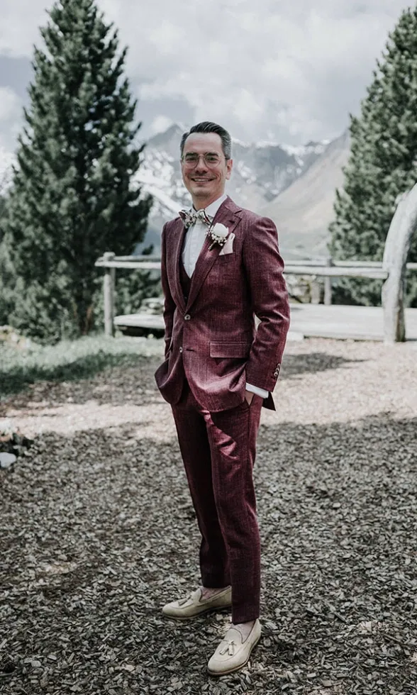 Bräutigam im bordeauxfarbenen Hochzeitsanzug steht entspannt im Freien, im Hintergrund eine alpine Berglandschaft mit Bäumen und Holzgeländer.