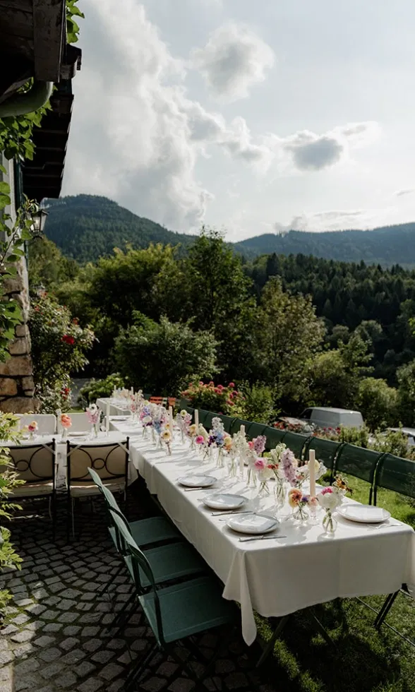 Lange Hochzeitstafel im Freien mit weißer Tischdecke, floraler Dekoration und Blick auf grüne Berge.