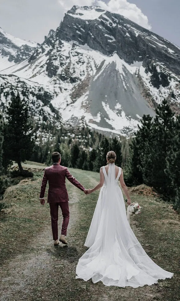 Braut und Bräutigam gehen Hand in Hand einen Weg durch die Alpen, von hinten fotografiert, mit schneebedecktem Bergmassiv vor sich.