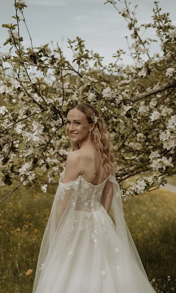 Braut steht vor einem blühenden Baum und blickt lächelnd über die Schulter, ihr Kleid weht leicht im Wind.