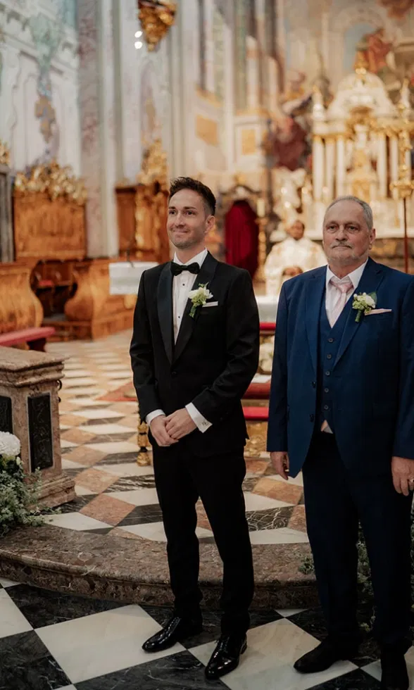 Bräutigam wartet gemeinsam mit einem Familienmitglied am Altar in der Kirche, festlich gekleidet vor kunstvoller Kulisse.